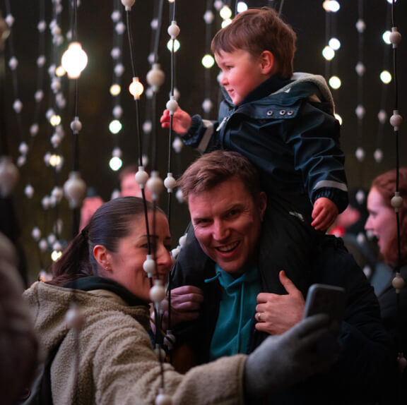 Two adults, one with a toddler on thier shoulders, taking a selfie surrounded by lights