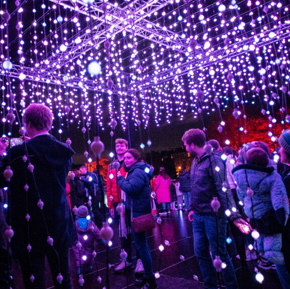 A large group of people walking through a maze of illuminated strings hanging from above